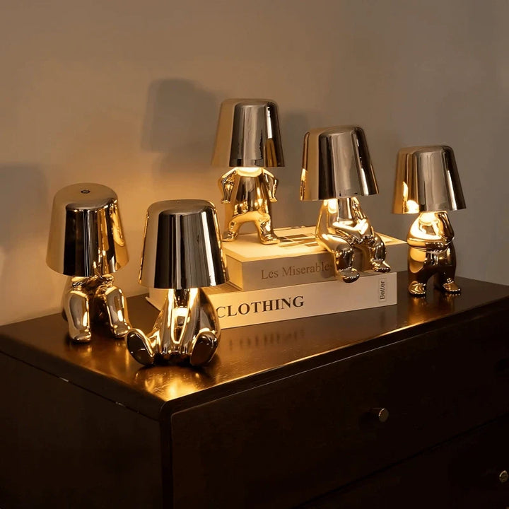 Designerska lampa stołowa LED siedzący człowiek z połyskującym wykończeniem w złotym i srebrnym kolorze na ciemnej komodzie